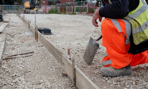 groundworker-orange-safety-hi-vis-trousers-fixing-timber-along-string-line-with-steel-pin-form-kerb-riser-straight-edge-tarmac-road-surface-during-new-road-construction